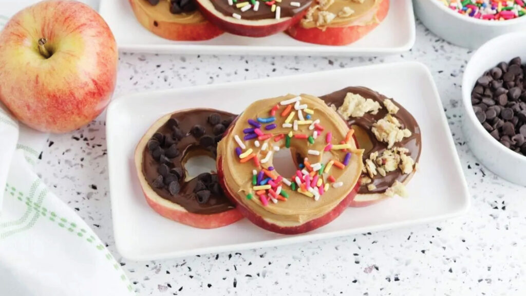 Apple slices topped with chocolate, peanut butter, sprinkles, and chocolate chips on a white plate.