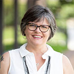 A smiling woman with short gray hair wearing glasses and a white sleeveless top, standing outdoors.
