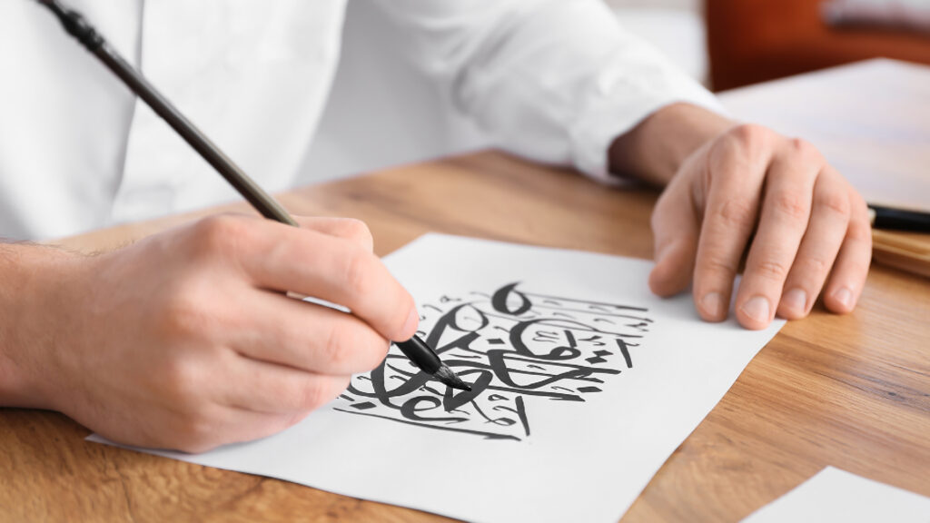 Person writing Arabic calligraphy with a black pen on white paper at a wooden table.