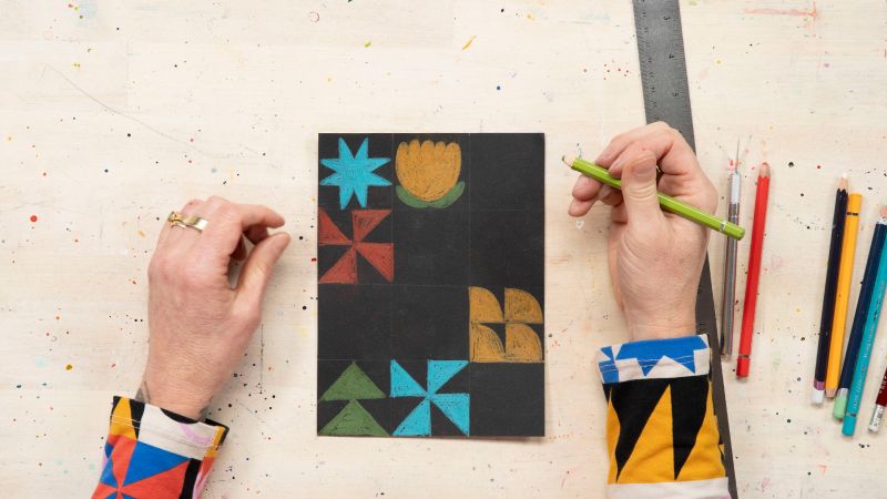 Person drawing colorful geometric shapes on black paper with pencils on a splattered surface.