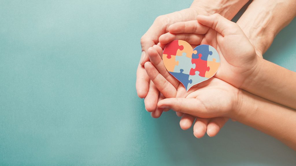 Hands holding a heart-shaped puzzle piece with multicolored segments against a blue background.