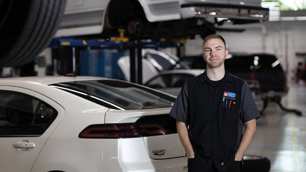 Auto mechanic stands beside a white car in a garage with vehicles on lifts in the background.