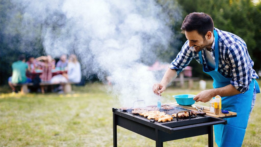 Man grilling food on a barbecue grill outdoors, with a group of people seated at a picnic table in the background.