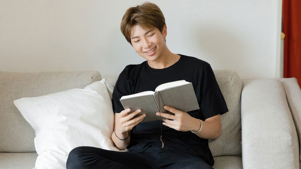 A person sitting on a couch, smiling while reading a book, dressed in a black shirt and pants.
