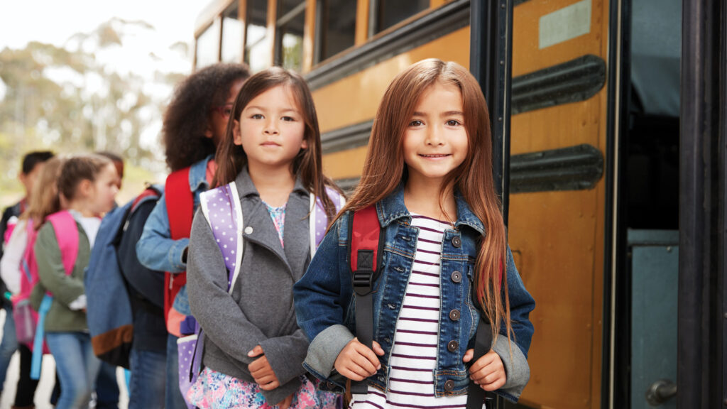 Children in a line, smiling, as they board a yellow school bus. They are wearing backpacks and casual clothing.