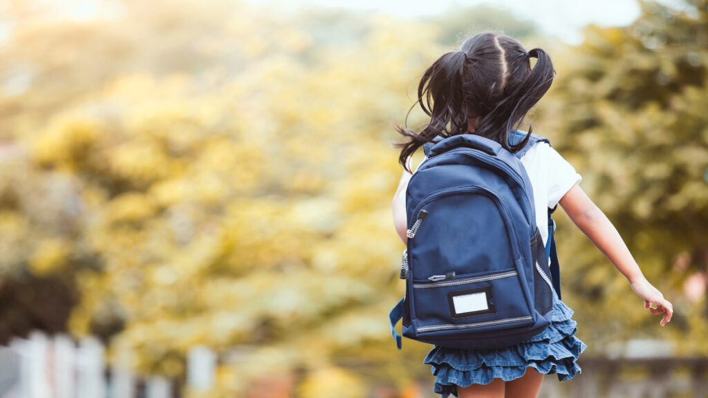 A girl with a blue backpack and pigtails walks through a park with yellow foliage, captured from behind.