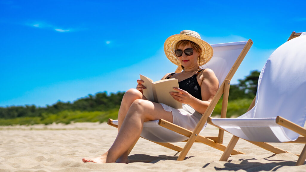 Woman in a sunhat and sunglasses reading a book while relaxing on a beach chair under a clear blue sky.