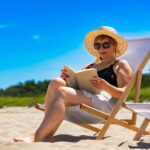 Woman in a sunhat and sunglasses reading a book while relaxing on a beach chair under a clear blue sky.
