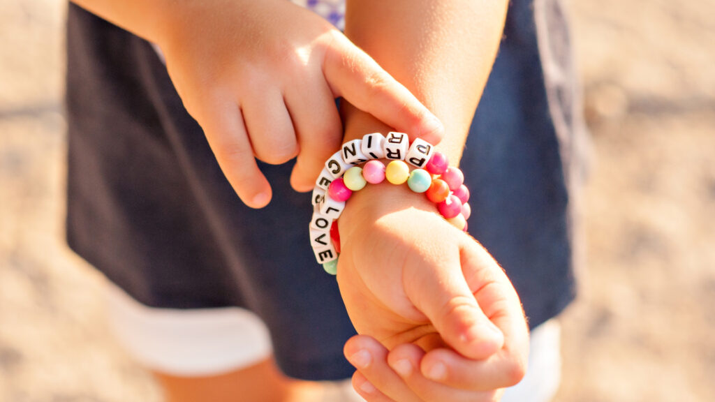 A child wears colorful beaded bracelets with words, including "PEACE," "LOVE," and "FRIEND," on their wrist.