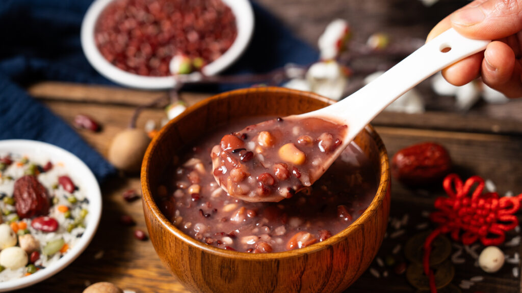 A hand holds a spoon of red bean porridge above a wooden bowl, with nuts and beans around.