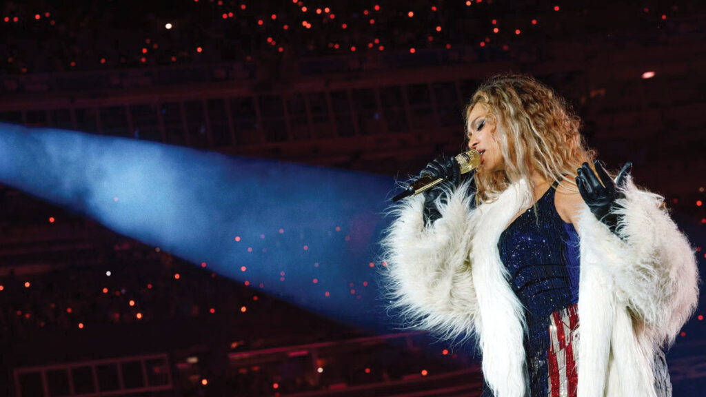 Singer in a white fur coat performing on stage with a spotlight and red lights in the background.