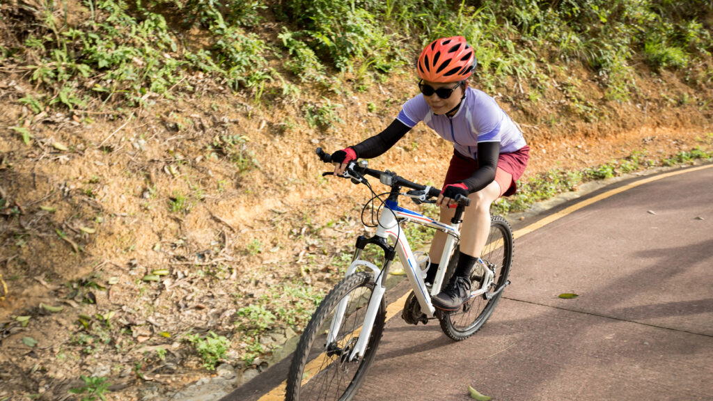 Person wearing a helmet and sunglasses rides a bicycle uphill on a paved road beside greenery.