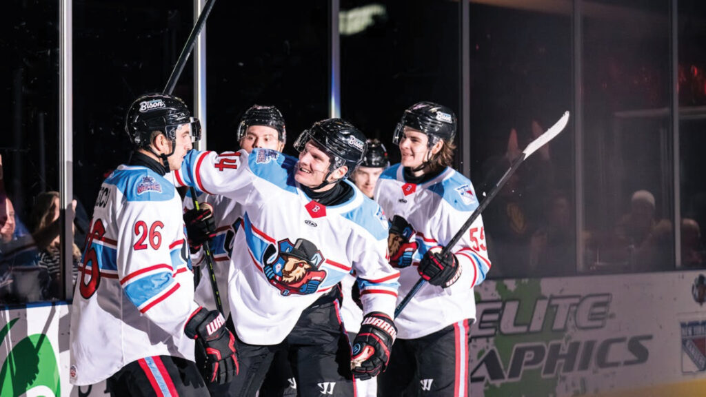 Four ice hockey players in white jerseys celebrate together on the rink during a game.