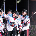 Four ice hockey players in white jerseys celebrate together on the rink during a game.
