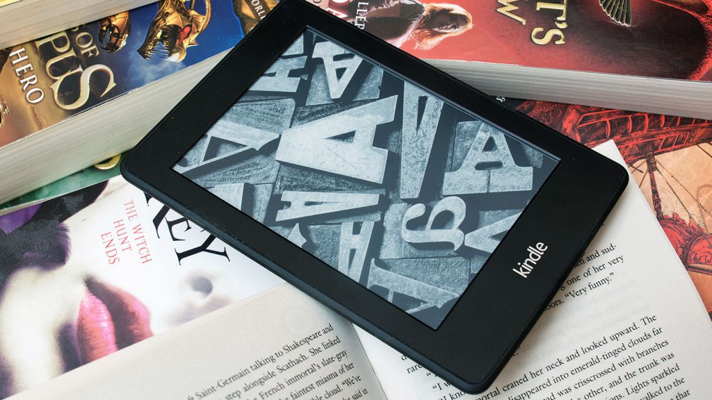 A Kindle e-reader is placed on a stack of open and closed books with colorful covers.