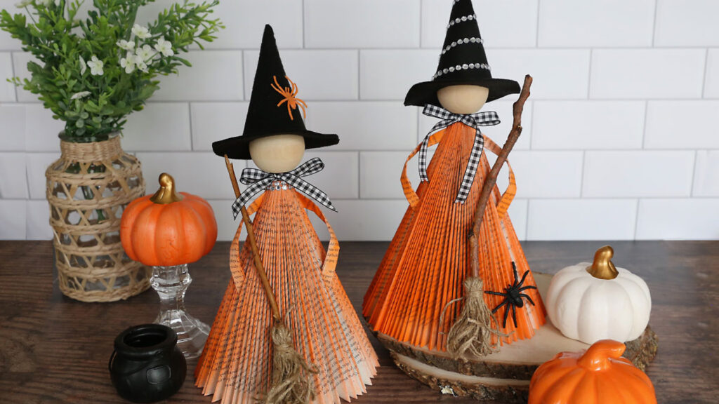 Two handmade witch dolls with orange dresses stand among pumpkins and Halloween decorations on a wooden surface.