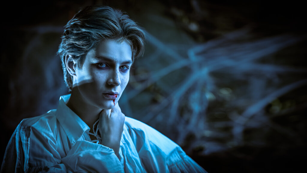 Person with pale skin, red lips, and fangs in a white shirt posing in eerie blue lighting.