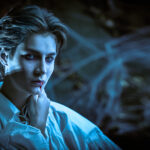 Person with pale skin, red lips, and fangs in a white shirt posing in eerie blue lighting.