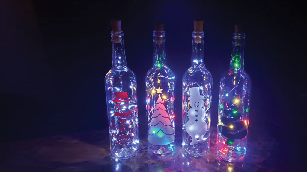 Four glass bottles with corks, filled with string lights and festive Christmas decorations inside each bottle.