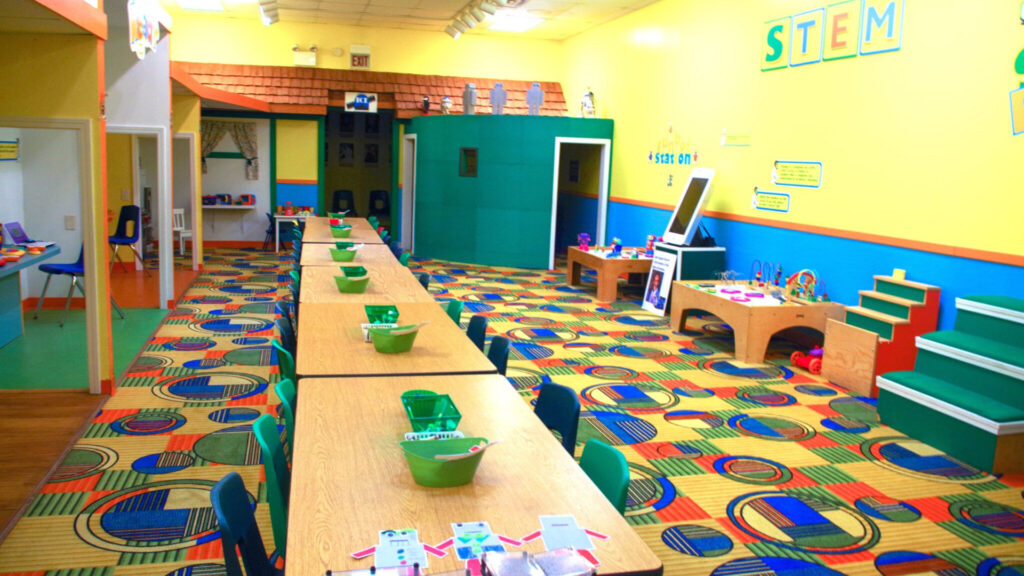 A colorful classroom with a long table, chairs, activity stations, and STEM learning decorations on the wall.