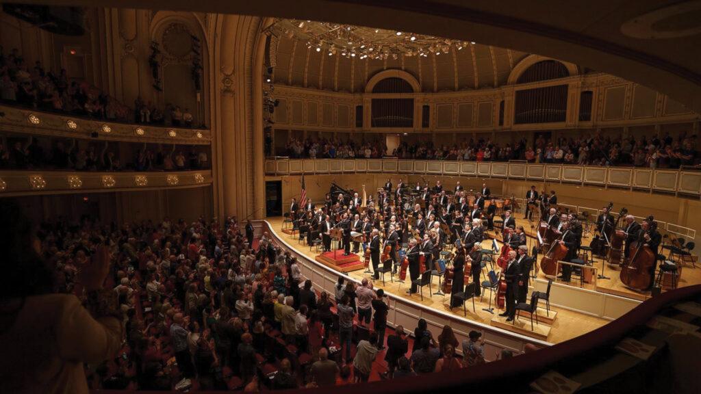 Orchestra on stage performing in a grand concert hall with a large audience applauding.