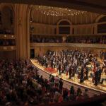 Orchestra on stage performing in a grand concert hall with a large audience applauding.