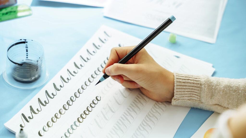 A person practices calligraphy on a worksheet with a black pen, surrounded by other calligraphy materials.