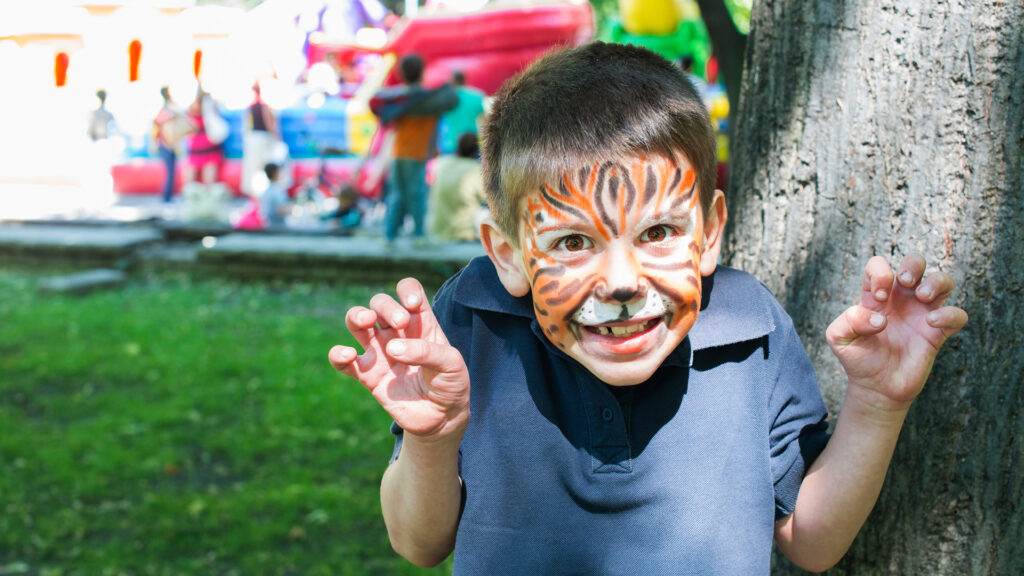 A child with face paint resembling a tiger playfully poses with claws up in a park with a bouncy house in the background.