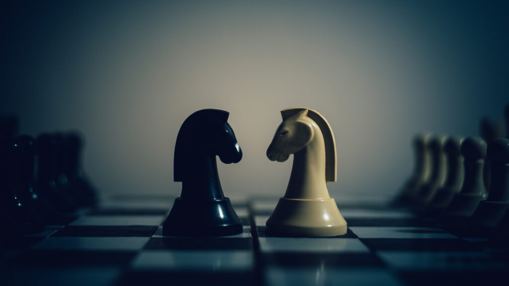 A black and a white chess knight face each other on a chessboard in a dimly lit, high-contrast close-up shot.