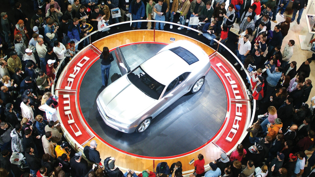 A silver car is displayed on a circular platform surrounded by a crowd at an auto show.