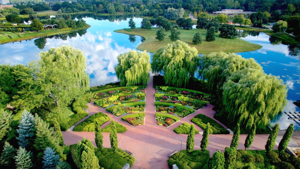 Aerial view of a landscaped garden with colorful flower beds, trees, and a reflective lake in the background.