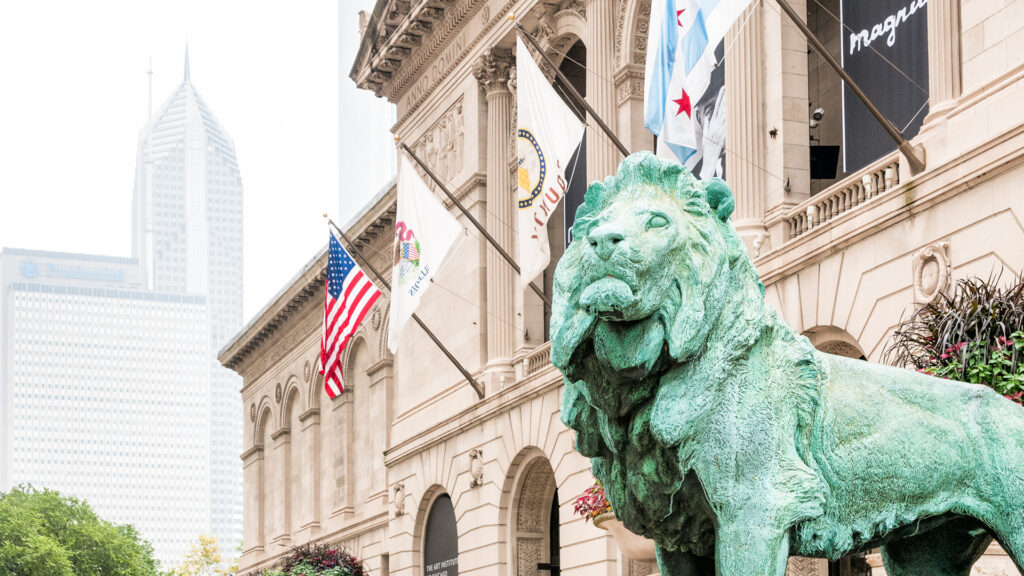 A bronze lion statue stands outside a museum with flags and a city skyscraper in the background.