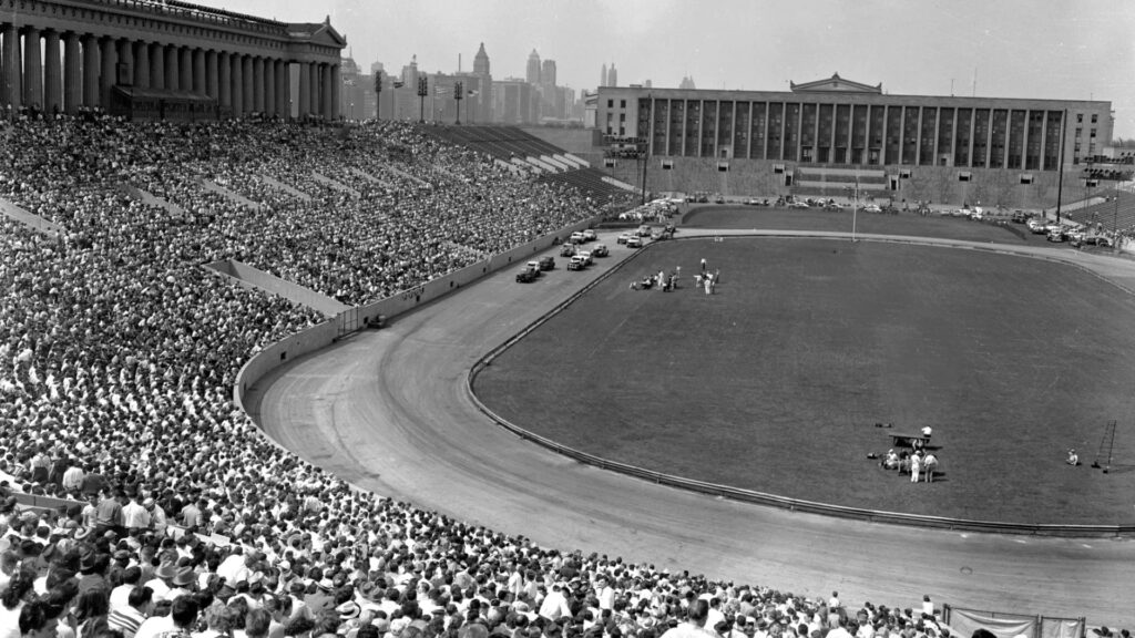 A large crowd fills a historic stadium with a running track and city skyline in the background.
