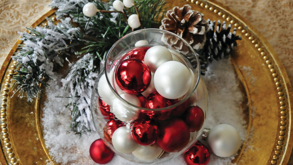 Glass vase with red and white ornaments on a gold plate, surrounded by pine cones and snowy branches.