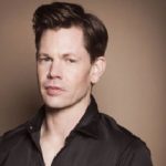 A man with short brown hair wearing a black shirt stands against a beige background.