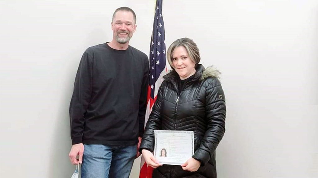 Two people stand smiling; one holds a certificate in front of a U.S. flag.