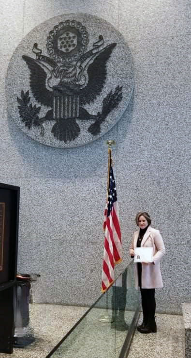 A person holds a certificate, standing beside a U.S. flag in front of a large U.S. seal on a gray wall.