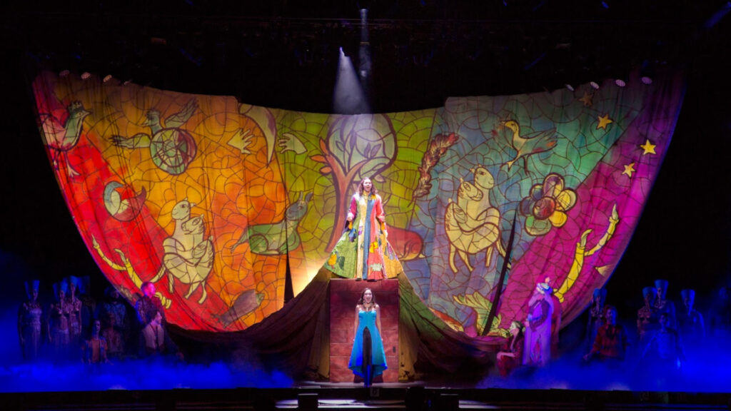 Vibrant stage set with colorful stained glass wings backdrop, performers in elaborate costumes, and dramatic lighting.