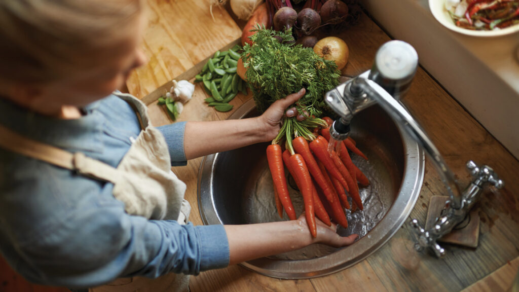 Person rinsing a bunch of fresh carrots under a kitchen faucet with other vegetables nearby on the counter.