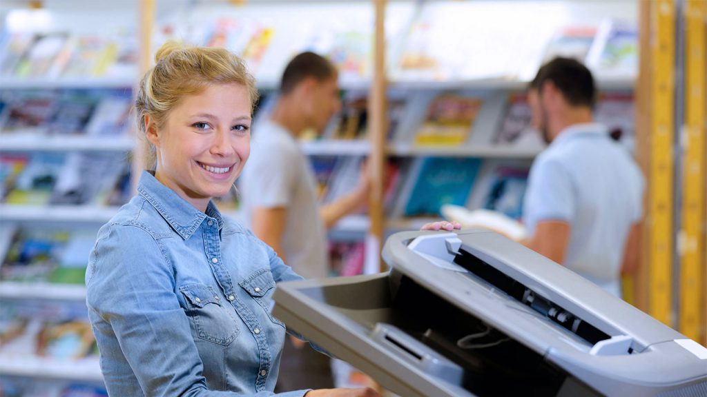 A woman smiles while using a photocopier in a library with two men browsing books in the background.