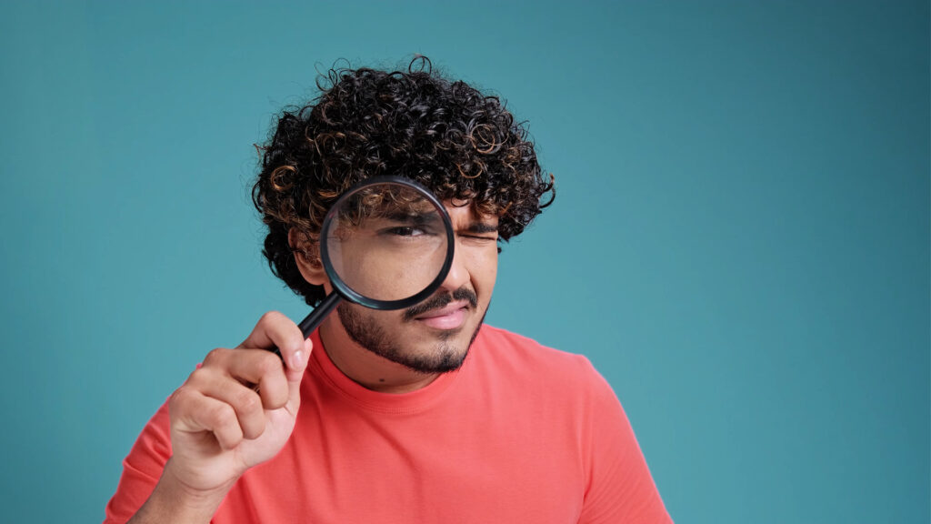 A man with curly hair in a red shirt holding a magnifying glass to his eye against a teal background.