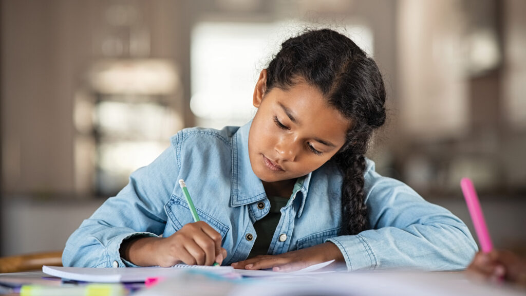 A young girl with braided hair writes in a notebook while sitting at a table, focused on her work.