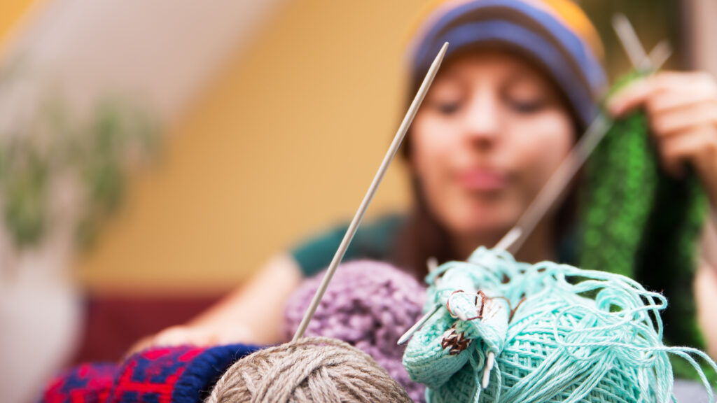 A person in a colorful hat holds knitting needles and yarn, with balls of yarn in the foreground.