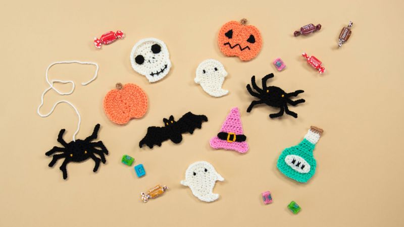 Crocheted Halloween items, including a pumpkin, ghost, skull, bat, witch hat, and candy, are spread on a beige background.