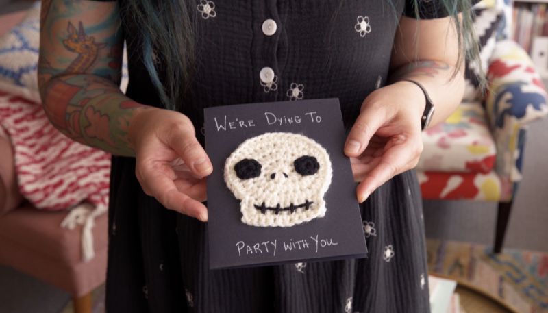 Person holding a card with a crocheted skull and the text "We’re Dying to Party With You.