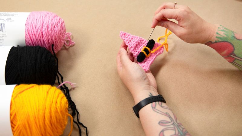 Hands crocheting with pink yarn, surrounded by balls of yellow, black, and pink yarn on a beige surface.