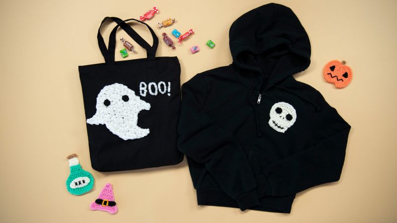 Halloween-themed tote bag with ghost and "Boo!" text, black hoodie with a skull, candy, and knitted decorations.