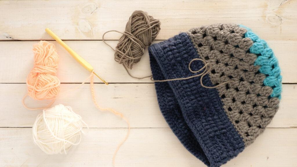 Crocheted hat with blue, grey, and teal yarn beside balls of yarn and a crochet hook on a wooden surface.