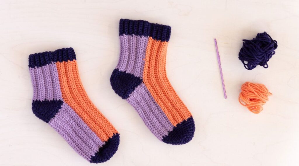 Crocheted socks in purple, orange, and navy, with a crochet hook and two yarn balls on a white background.