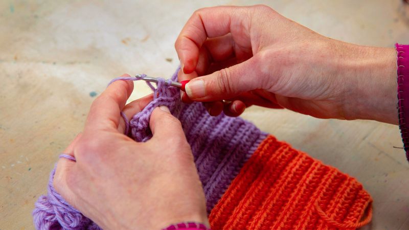 Close-up of hands crocheting with purple yarn, creating a piece that transitions into orange.